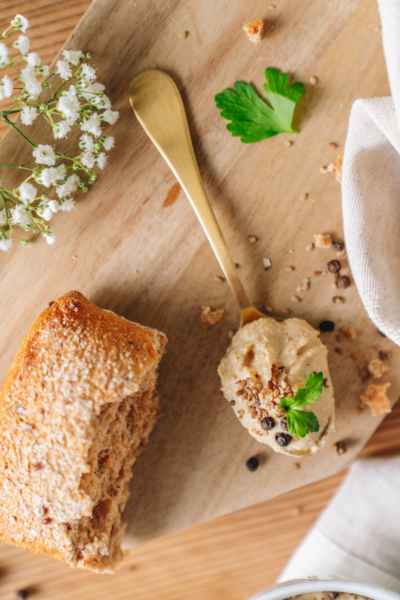 Bio-Hummus auf einem Löffel, der neben einem halben Weckerl auf einem Holzbrett liegt.