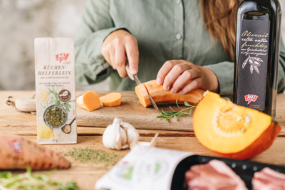 Jemand schneidet eine Bio-Süßkartoffel auf einem Schneidebrett, umgeben von Produkten von Ja! Natürlich.