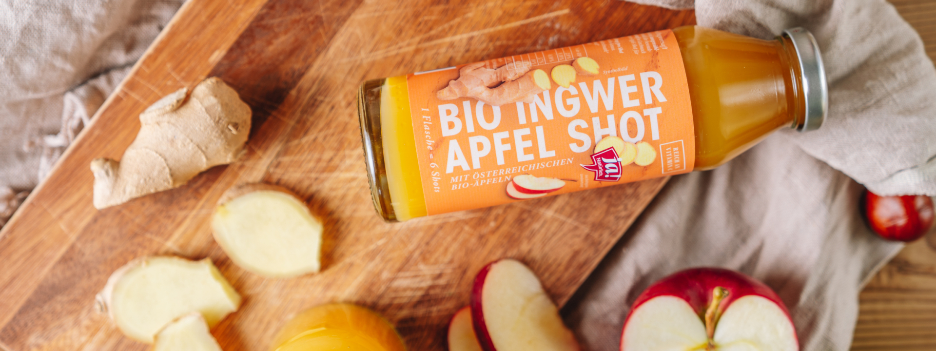 Auf einem Brett liegt eine Flasche Ja! Natürlich Bio-Ingwer-Apfel-Shot und die Zutaten.