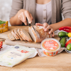 Ja! Natürlich Bio-Gemüseaufstrich und -Käse. Im Hintergrund schneidet jemand einen Laib Brot.