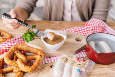 Jemand taucht eine Weißwurst in Senf, daneben liegt eine Brezel und eine Packung Bio-Weißwürste von Ja! Natürlich.
