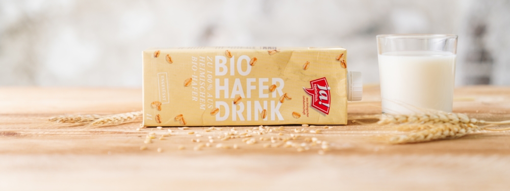 Ein Karton Ja! Natürlich Bio-Hafer Drink mit einem Glas gefüllt mit dem Hafer Drink daneben.