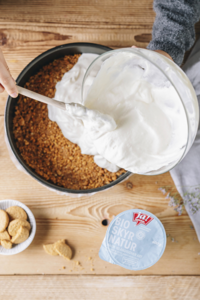Jemand gibt eine Masse aus Ja! Natürlich Bio-Skyr in eine Kuchenform mit Keksboden.