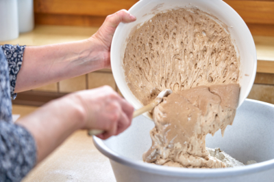 Jemand gibt Sauerteig mit einem Kochlöffel in eine Schüssel.