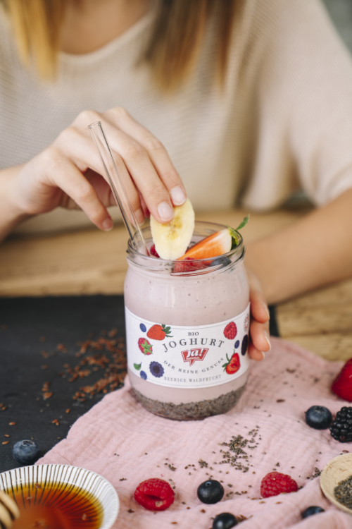 Jemand legt Bio-Früchte in ein Glas Ja! Natürlich Bio-Joghurt Beerige Waldfrucht.
