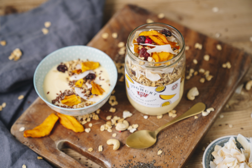 Ja! Natürlich Bio-Joghurt mit Granola, Trockenfrüchten und Mangoscheiben auf einem Holzbrett.