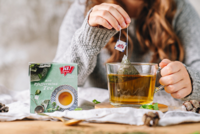 Jemand macht einen Ja! Natürlich Bio-Pfefferminztee in einer Tasse. Eine Packung des Tees steht daneben.