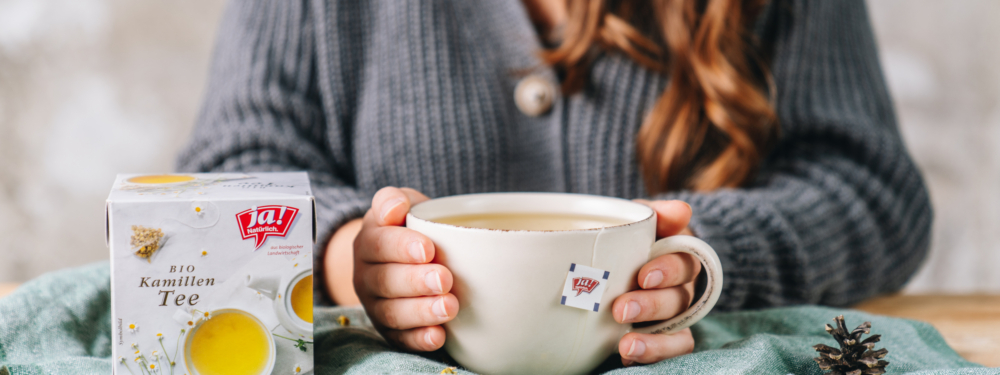 Eine Packung Ja! Natürlich Bio-Kamillen Tee und jemand wärmt sich die Hände an einer Tasse.