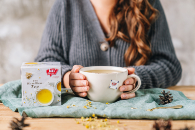 Eine Packung Ja! Natürlich Bio-Kamillen Tee und jemand wärmt sich die Hände an einer Tasse.