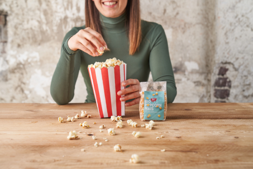 Jemand isst Popcorn, davor eine Packung Ja! Natürlich Bio-Popcorn-Mais.