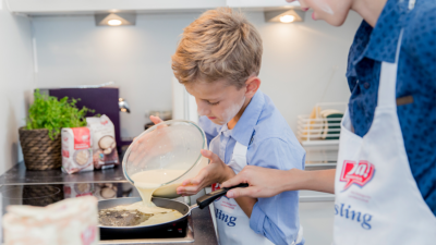 Kind schüttet Teig in Pfanne und Erwachsener steht daneben
