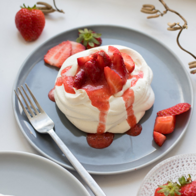 Pavlova auf einem Teller mit Bio-Erdbeeren und Sauce, daneben eine Gabel und ganze Erdbeeren.