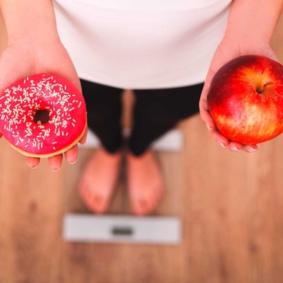 Person auf einer Waage, mit einem Donut und einem Apfel in der Hand.