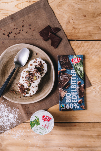 Ein Teller mit Ziegenjoghurt-Parfait garniert mit dunkler Schokolade. Daneben liegt eine Ja! Natürlich Bio-Edelbitterschokolade und ein Becher Ziegenjoghurt aus Heumilch.