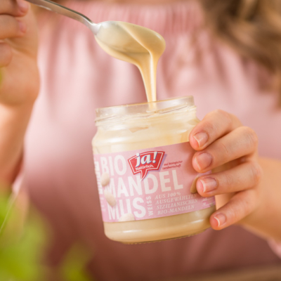 Jemand nimmt einen Löffel voll Ja! Natürlich Bio-Mandelmus aus dem Glas.