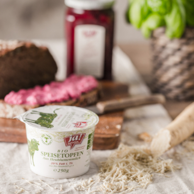 Becher Ja! Natürlich Bio-Speisetopfen und im Hintergrund eine Scheibe Brot mit Topfenaufstrich.