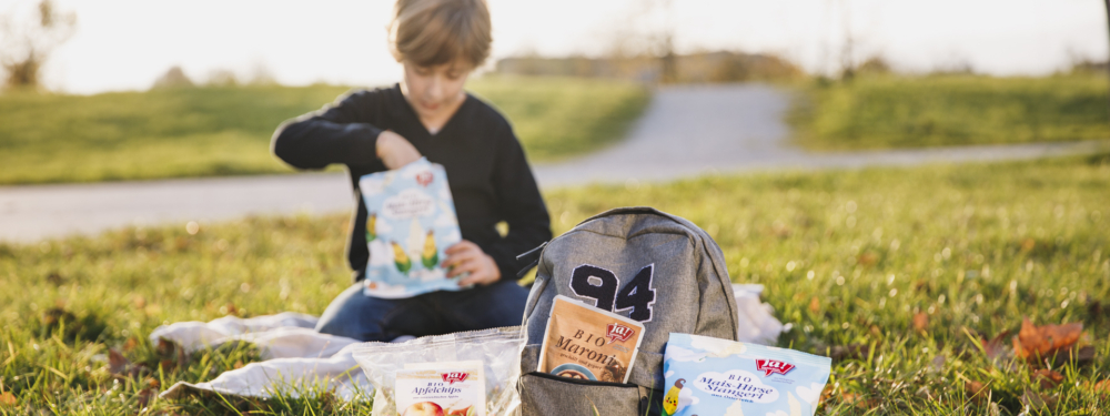 Ein Kind isst Ja! Natürlich Bio-Mais-Hirse-Stangerl auf einer Picknickdecke mit mehr Produkten von Ja! Natürlich.
