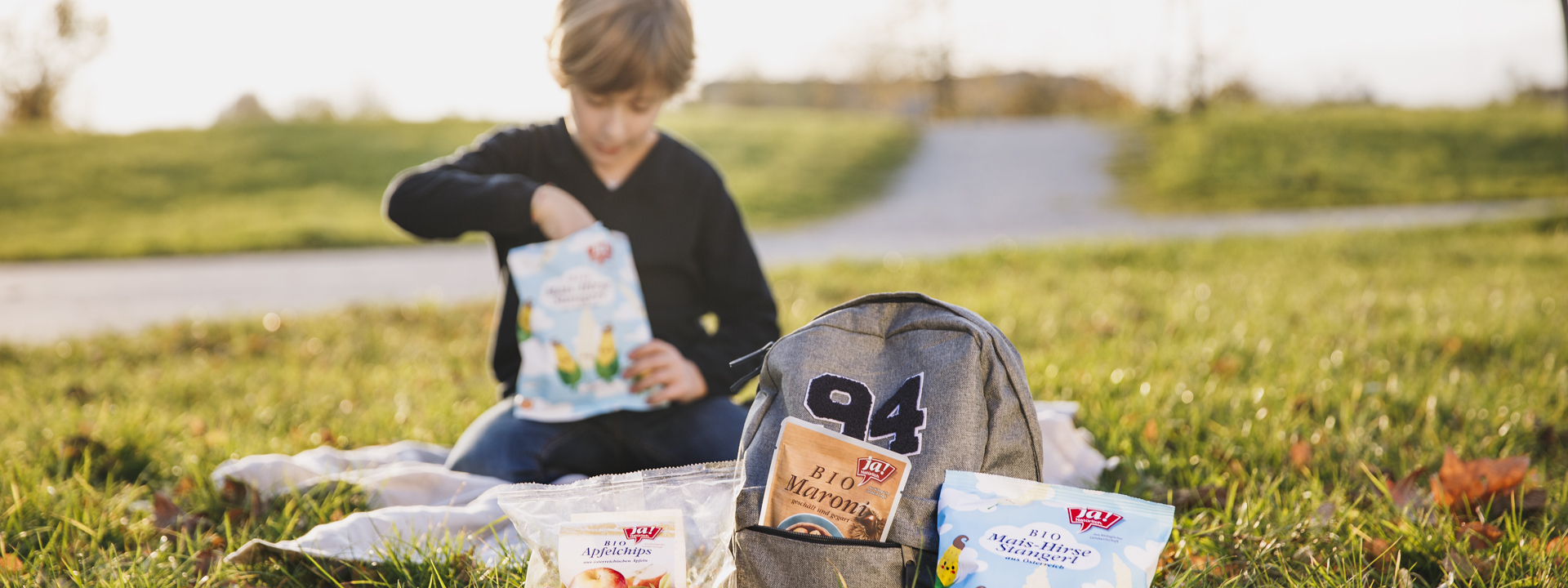 Ein Kind isst Ja! Natürlich Bio-Mais-Hirse-Stangerl auf einer Picknickdecke mit mehr Produkten von Ja! Natürlich.