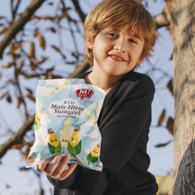 Kind hält Bio-Mais-Hirse-Stangerl von Ja! Natürlich in die Kamera.