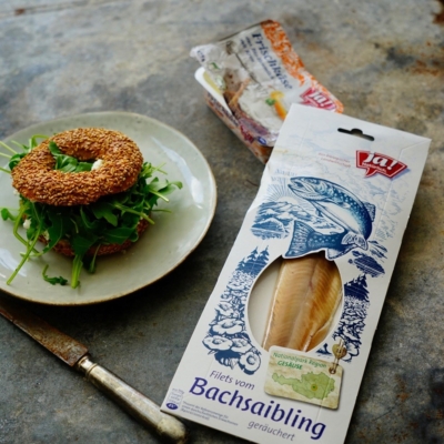 Ein Teller mit einem gefüllten Bagel und einer Packung Ja! Natürlich Bio-Bachsaibling.
