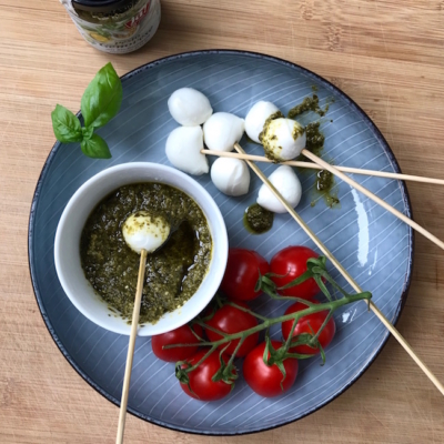 Auf einem Teller liegen Spieße mit Mozzarella, eine Bio-Paradeiserrebe und eine Schale mit grünem Pesto.