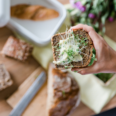 Jemand hält eine Scheibe Bio-Brot mit Butter und Sprossen über einen Tisch, auf dem Brote liegen.