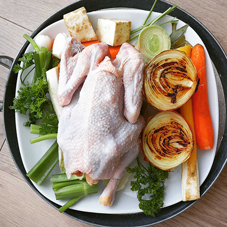 Auf einem Teller liegt ein ganzes rohes Hendl inmitten von Bio-Suppengemüse.