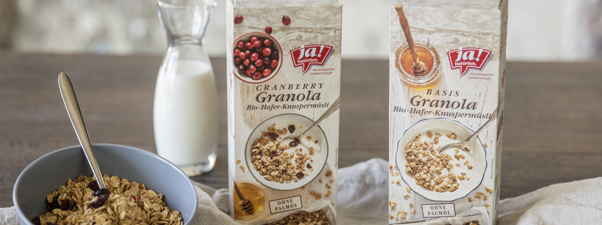 2 Packungen Ja! Natürlich Bio-Granola auf einem Tisch, eine mit Cranberrys und eine Schale Müsli davor.