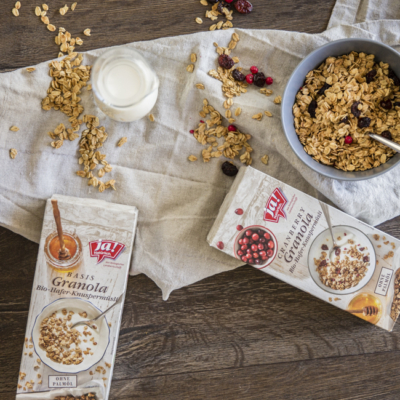 2 Packungen Ja! Natürlich Bio-Granola auf einem Tisch, eine mit Cranberrys und eine Schale Müsli davor.