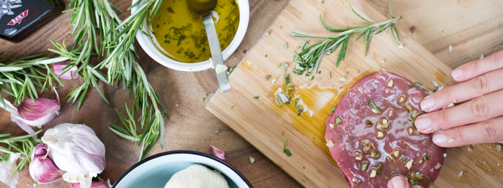 Jemand reibt Ja! Natürlich Bio-Olivenöl auf Bio-Fleisch, mit Knoblauch und Rosmarin daneben.