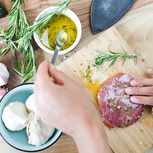 Würzen von rohem Bio-Fleisch mit Kräutern und Öl auf einem Schneidebrett, mit Knoblauch und Rosmarin daneben.