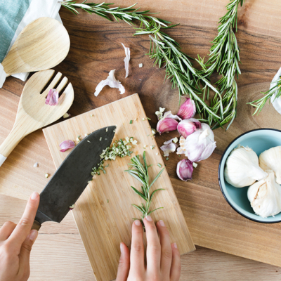 Jemand hackt Bio-Rosmarin auf ein Holzbrett, daneben liegen Knoblauch und Küchenwerkzeug bereit.